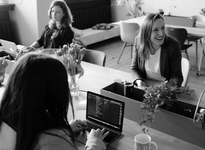 Three women in a workplace setting working on their laptops and conversing